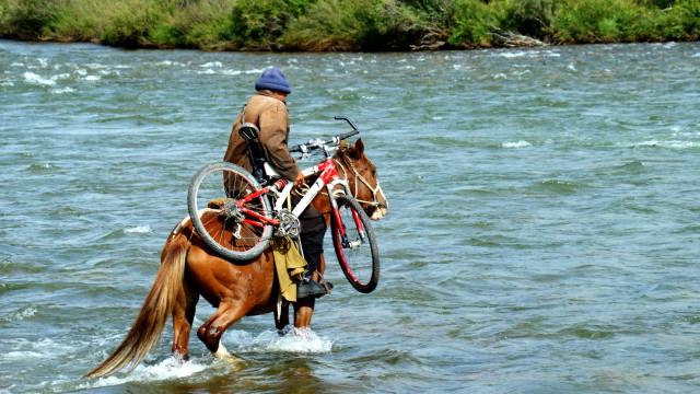 Man on horseback carrying a bicycle while attempting to cross a river.